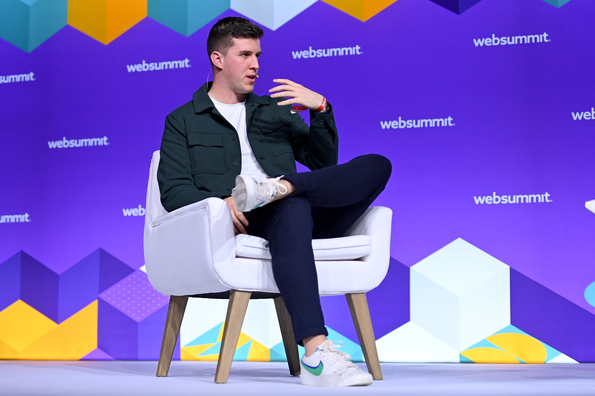Photograph of a person (Ciarán O'Mara, co-founder and CTO of Protex AI) speaking on stage at Deep Tech at Web Summit. They are sitting on a chair with one leg crossed over their other knee. They are gesturing with their hand.