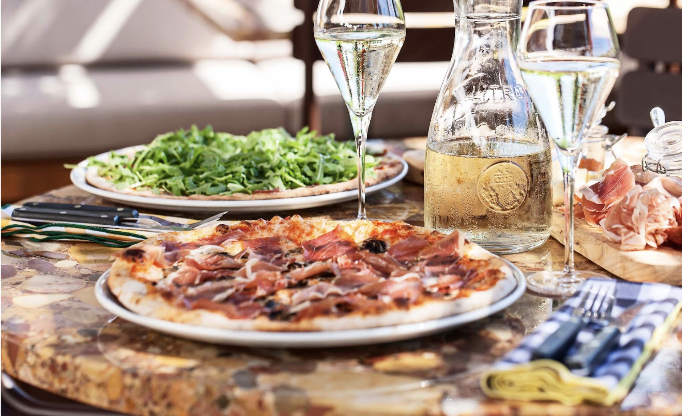 A pizza with olives and thinly sliced ham, a pizza piled high with rocket, two full wineglasses and a bottle of a water sit on a polished cork table.