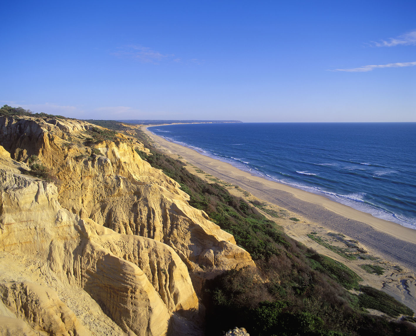 A long stretch of sandy coastline viewed from rocky hills. A thin sliver of grass and shrubs separates the hills from the beach. There are small white-capped waves in places. The sky is almost entirely clear of clouds.
