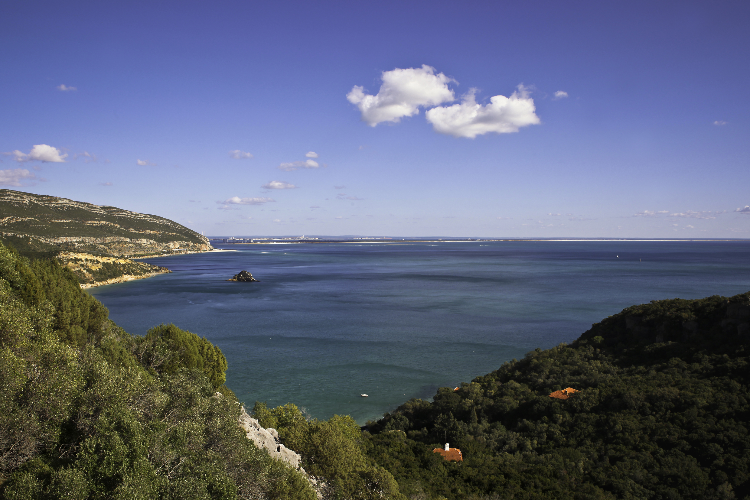 A bay, viewed from wooded hills. Small slivers of beaches are visible along the cliffs to the left of the bay. Red tiled roofs poke through the tree cover in two places. The water is clear, and there are a handful of small clouds in the sky.