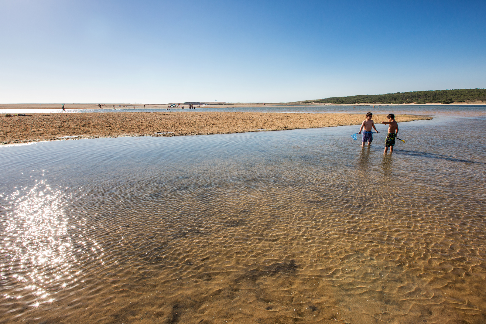 A thick band of shallow, clear water winds through a sandy expanse. Two young children stand in the water, talking to each other. One is holding a fishing net and the other is holding a spade. In the distance, a low hill covered in greenery. The sky is empty of clouds.