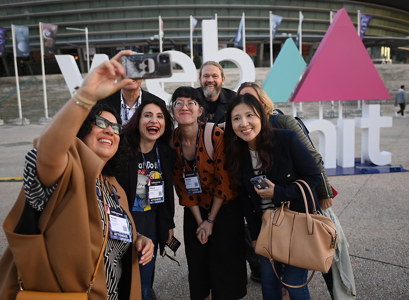 A diverse group of people pose for a selfie in front of a large, outdoor Web Summit sign. The people are smiling as they lean in for the photo. They are wearing lanyards and badges that read 'attendee'.