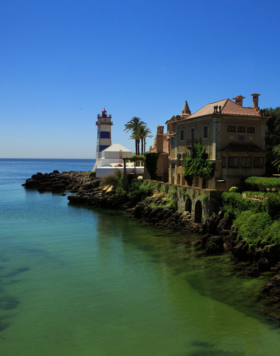 A spur of land juts into a clear ocean under cloudless daytime skies. On the spur stand a striped lighthouse, palm trees, and an old multi-storey building, which is covered in ivy. This is Cascais.