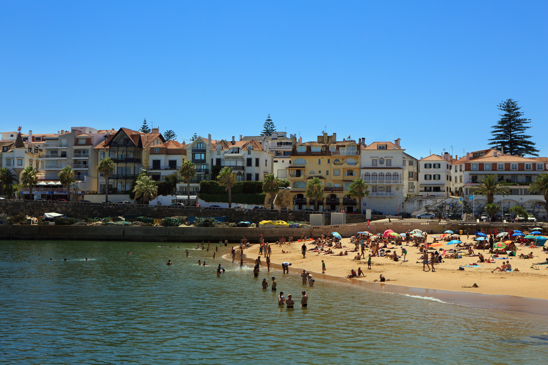 A row of multi-storey houses facing the sea. To the right, a sandy beach is nestled against a low sea wall. People lie on the beach under umbrellas while others walk the sands and swim in the calm waters.