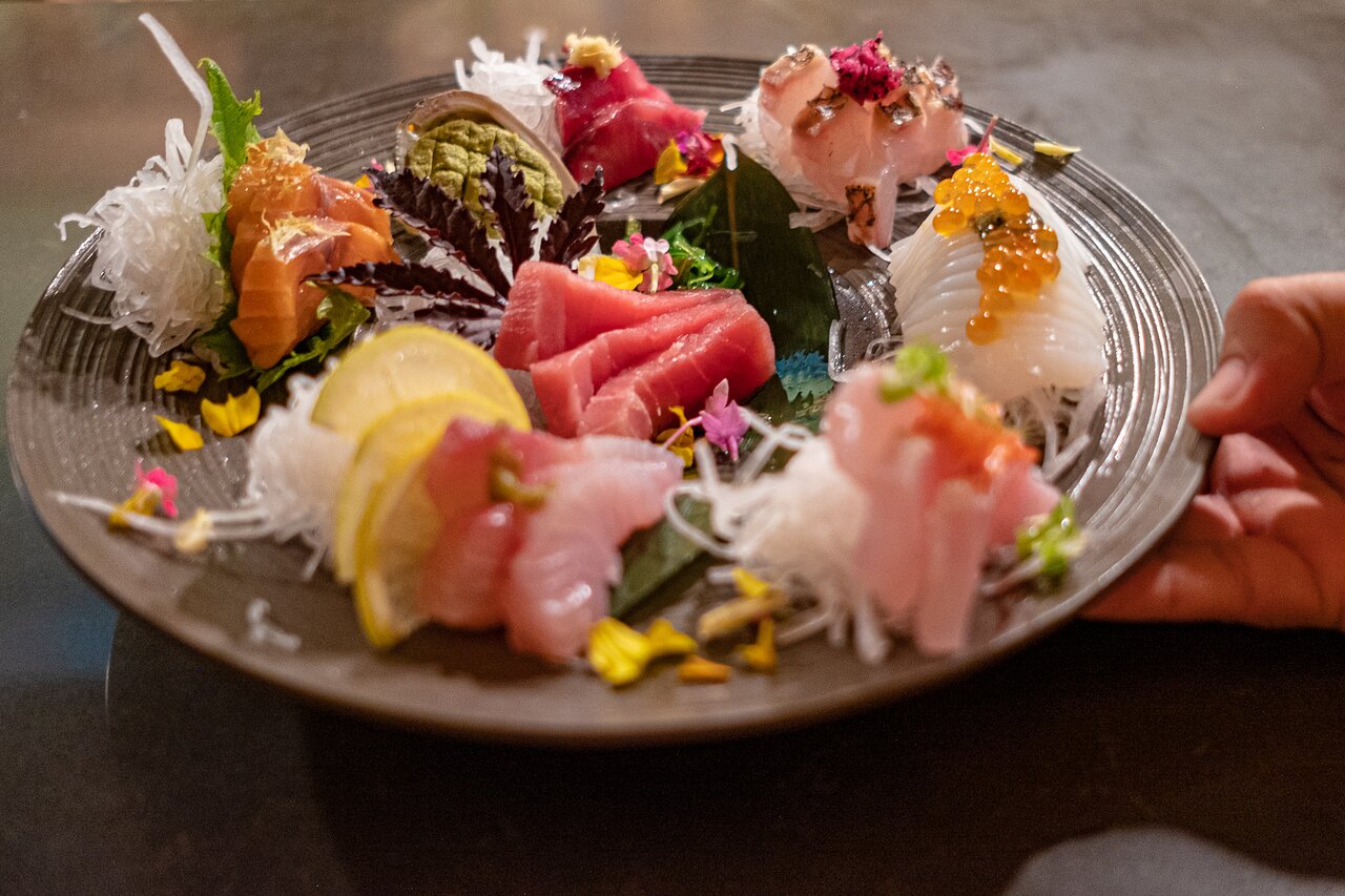 A hand holds a plate containing several varieties of sashimi, as well as rice noodles and vegetables.