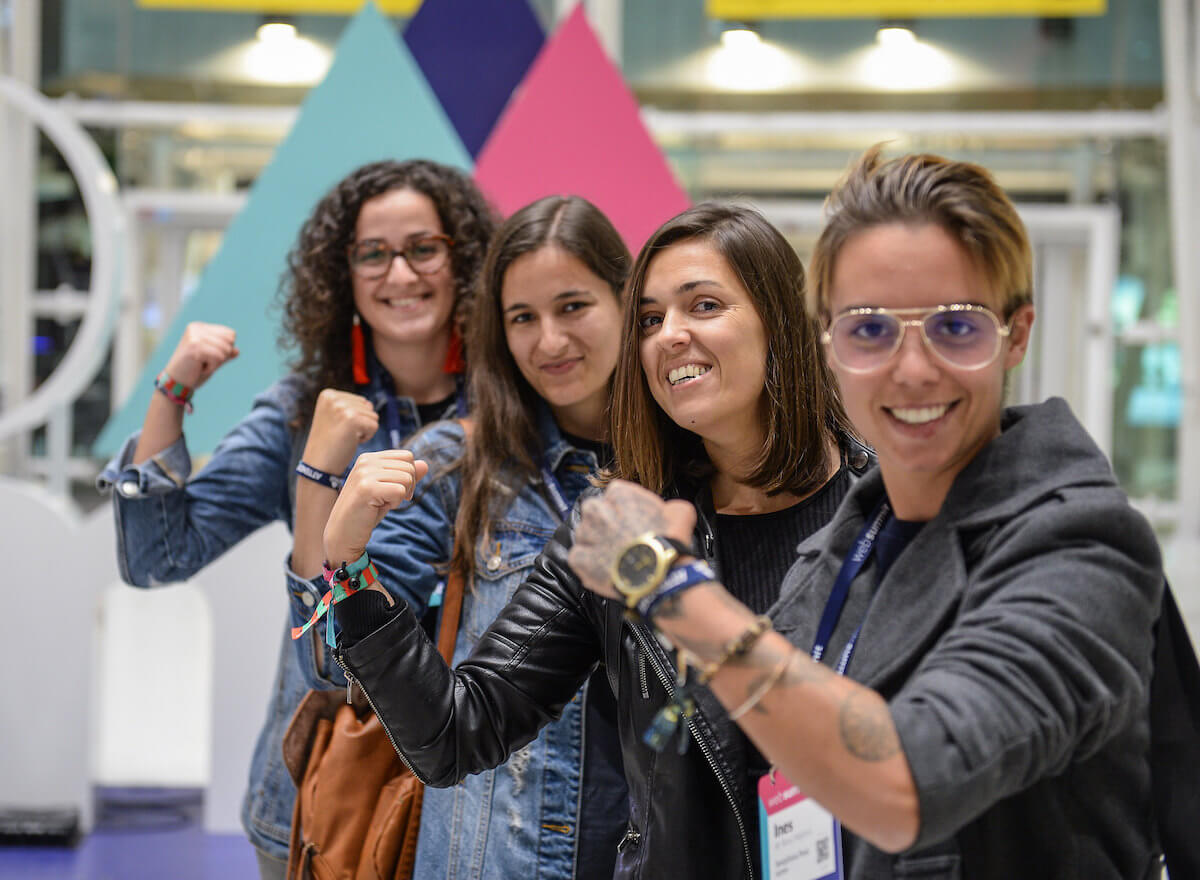 Four attendees showing their accreditation as they smile in front of the web summit branding at Lisbon airport
