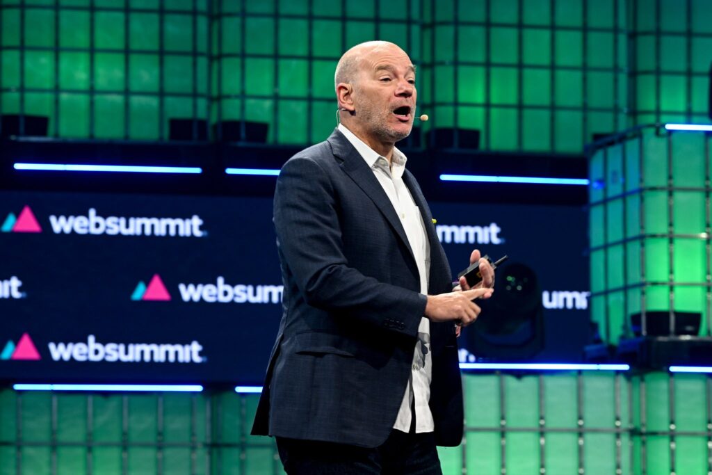 A person (MIT principal research scientist Andrew McAfee) walks across a stage. They are holding a presentation clicker in their left hand and wearing a headset mic. They appear to be speaking. The Web Summit logo is visible in several places behind them. This is Centre Stage during Day 1 of Web Summit 2023 at the Altice Arena in Lisbon, Portugal.