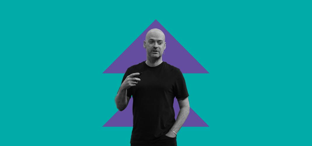 An image of a person (Christian Weedbrook, founder and CEO of Xanadu) wearing a t-shirt. They appear to be talking while gesturing with their hands. The background contains two arrow shapes, pointing upwards and stacked on top of each other.