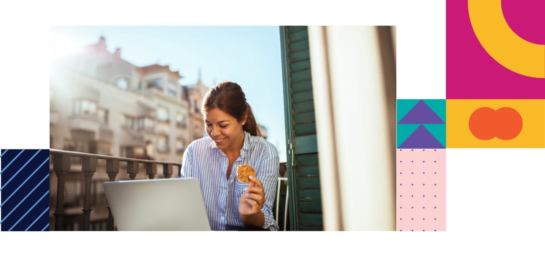 A woman in business attire sits on a balcony holding a cookie and smiling at her laptop