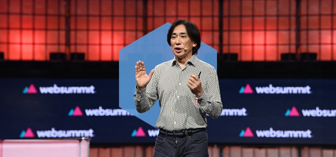 A person (Sega COO Shuji Utsumi) standing, pictured from the waist up. They are wearing a headset mic and gesturing emphatically with both hands. They appear to be speaking. The Web Summit logo is visible in several places behind them. This is Centre Stage at Web Summit.