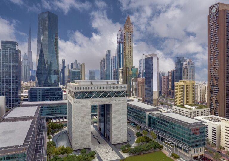 A modern cityscape boasting dozens of high-rise buildings, viewed from a high vantage point. In the foreground is a smaller multi-storey building in the shape of an arch. This arch-style building is surrounded by even lower multi-storey, glass-fronted buildings. This is Dubai.