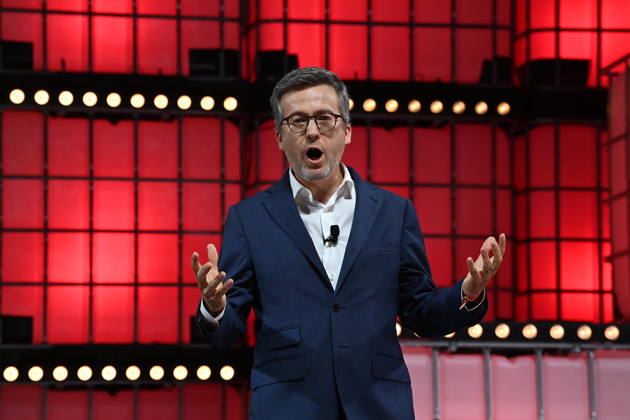 An image of Carlos Moedas, mayor of Lisbon. Carlos is looking directly at the camera and gesturing emphatically with both hands. Carlos is wearing what appears to be a small microphone attached to a shirt. Carlos appears to be speaking.