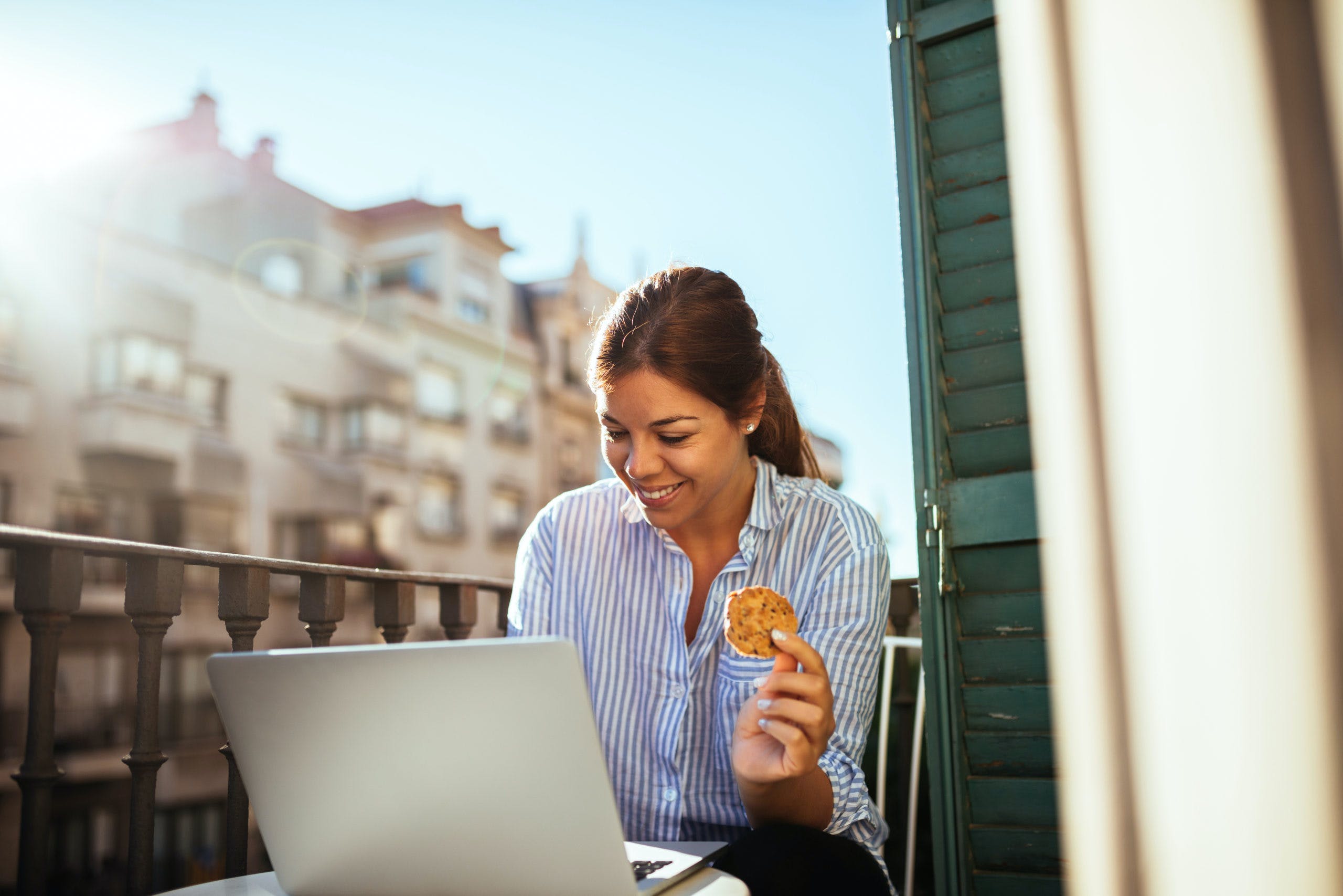 A woman in business attire sits on a balcony holding a cookie and smiling at her laptop