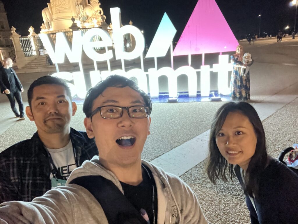 A selfie photograph of three people at Web Summit 2023. The person taking the selfie has their mouth open, and the other two people are smiling. In the background is a large, lit-up Web Summit logo.