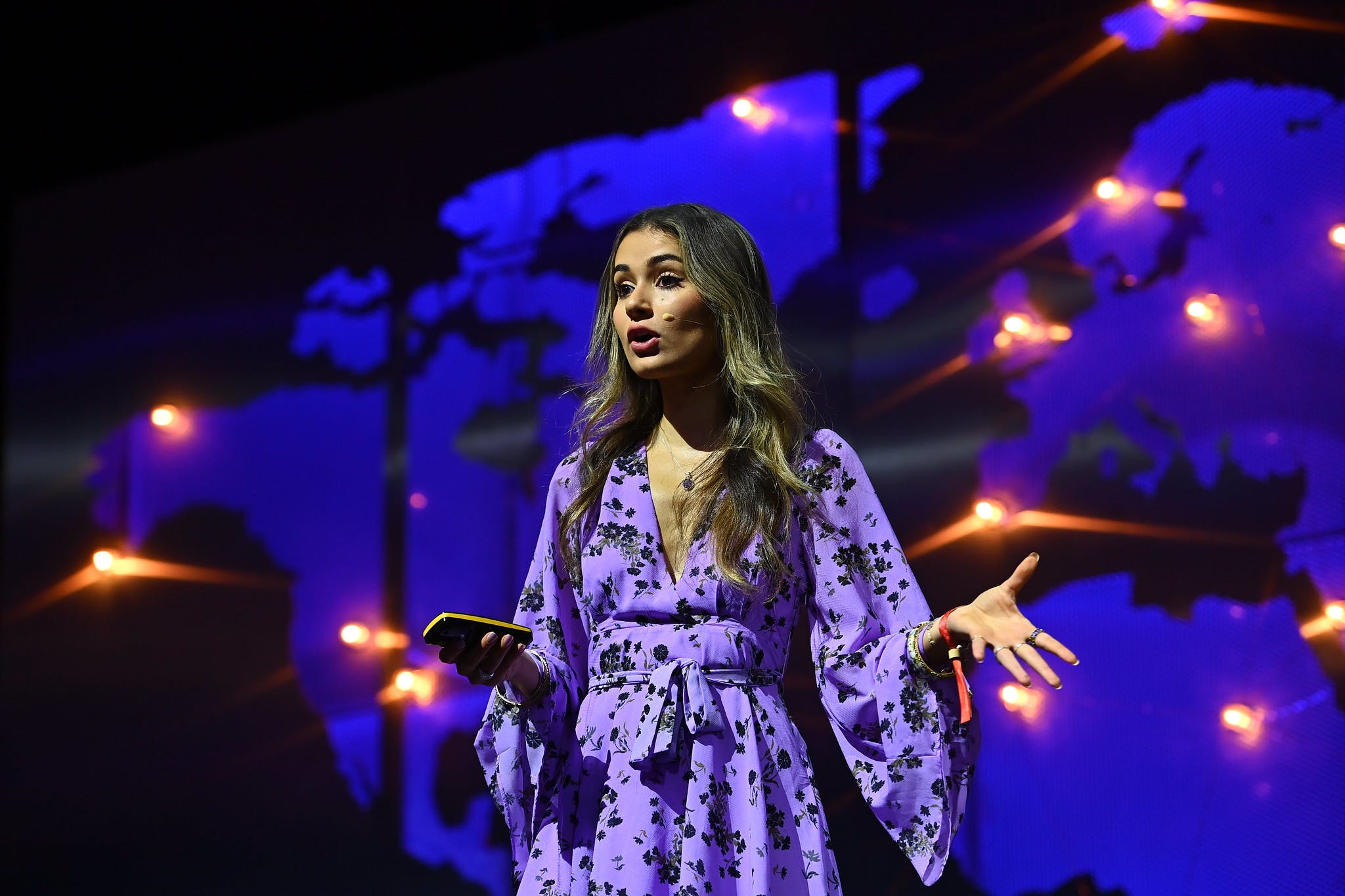 An image of Sophia Kianni, founder and executive director of Climate Cardinals. Sophia is standing on a stage and gesturing emphatically. Jess is holding what appears to be either a presentation clicker or a mobile phone, and appears to be talking. Jess is wearing a headset mic, and several lights emitting a warm glow are visible on the wall behind Sophia.