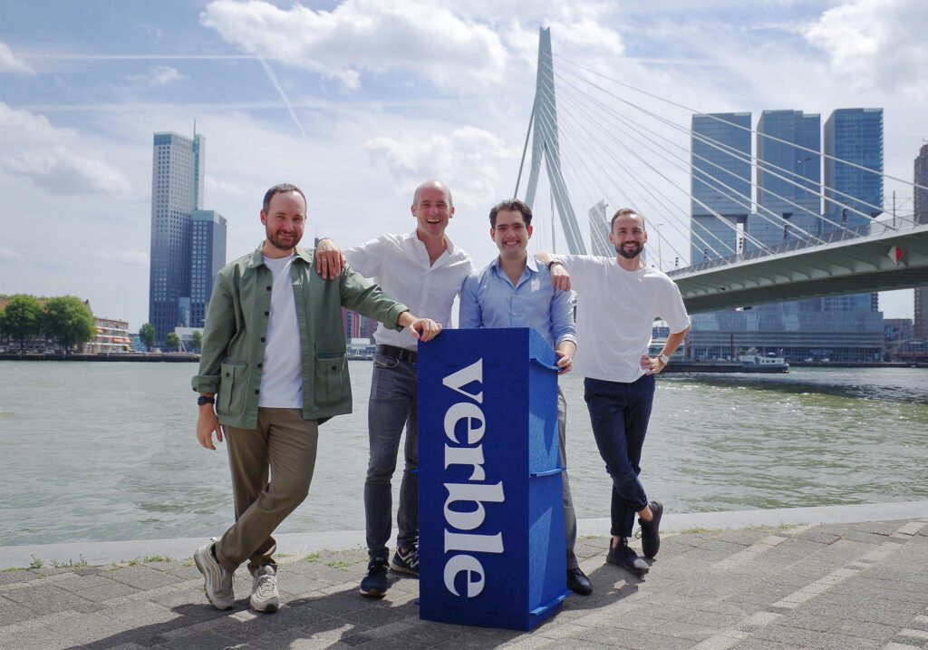 Four men are pictured wearing casual shirts and trousers stand around a sign that reads 'Verble'. In the background is a river with a suspended bridge and four skyscrapers towering above the city. It is a cloudy day.