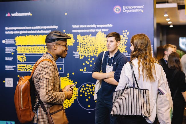 Three people stand in a circle in front of a wall with Entrepreneurs’ Organization written on it. The wall features survey questions, and stickers have been added to the wall to indicate responses to these questions. The three people appear to be in conversation.