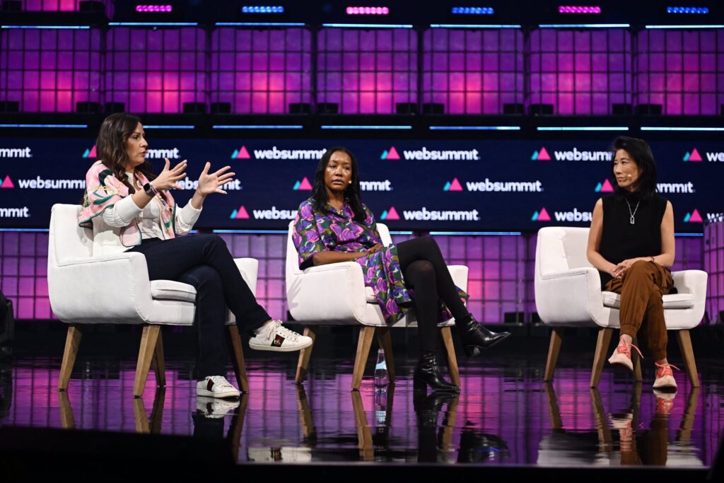 Three people sitting in chairs onstage. The person sitting on the left is speaking expressively, using their hands. Web Summit logos appear on the stage wall in the background.
