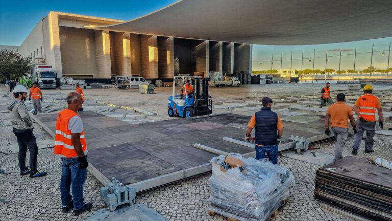 a building site, with several people wearing hard hats and hi-vis safety clothing. They are at work around a modular floor that's being built on top of cobbles. There is a large modern building in the background, and a forklift  is adding a section of floor. This is outside in Lisbon, Portugal.