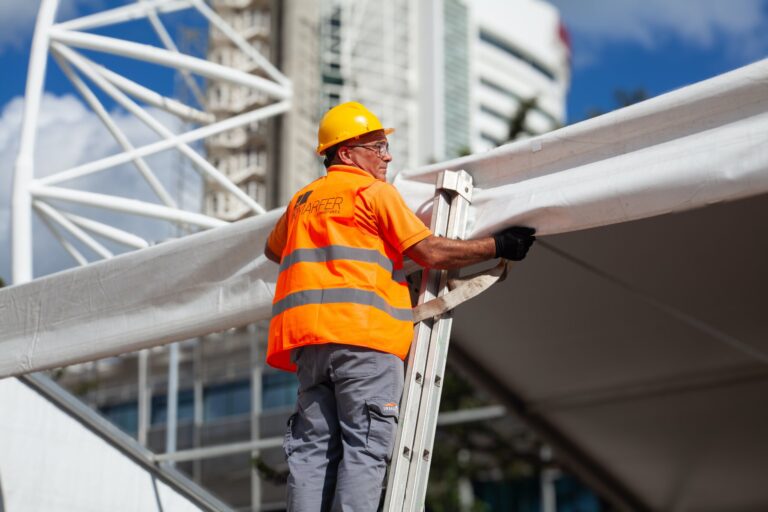 a person wearing a hard hat, cargo pants and a hi-vis safety vest stands atop a ladder that's leaning against a structure made of canvas and steel