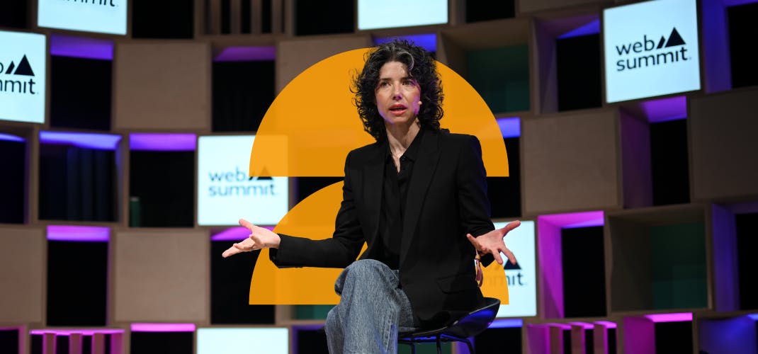 A person(Signal president Meredith Whittaker) sits on a high stool. The person is gesturing with both hands and is wearing a headset mic. They appear to be speaking. The Web Summit logo appears behind them on wooden boxes. This is Q&A stage during Day 1 of Web Summit 2023 at the Altice Arena in Lisbon, Portugal.