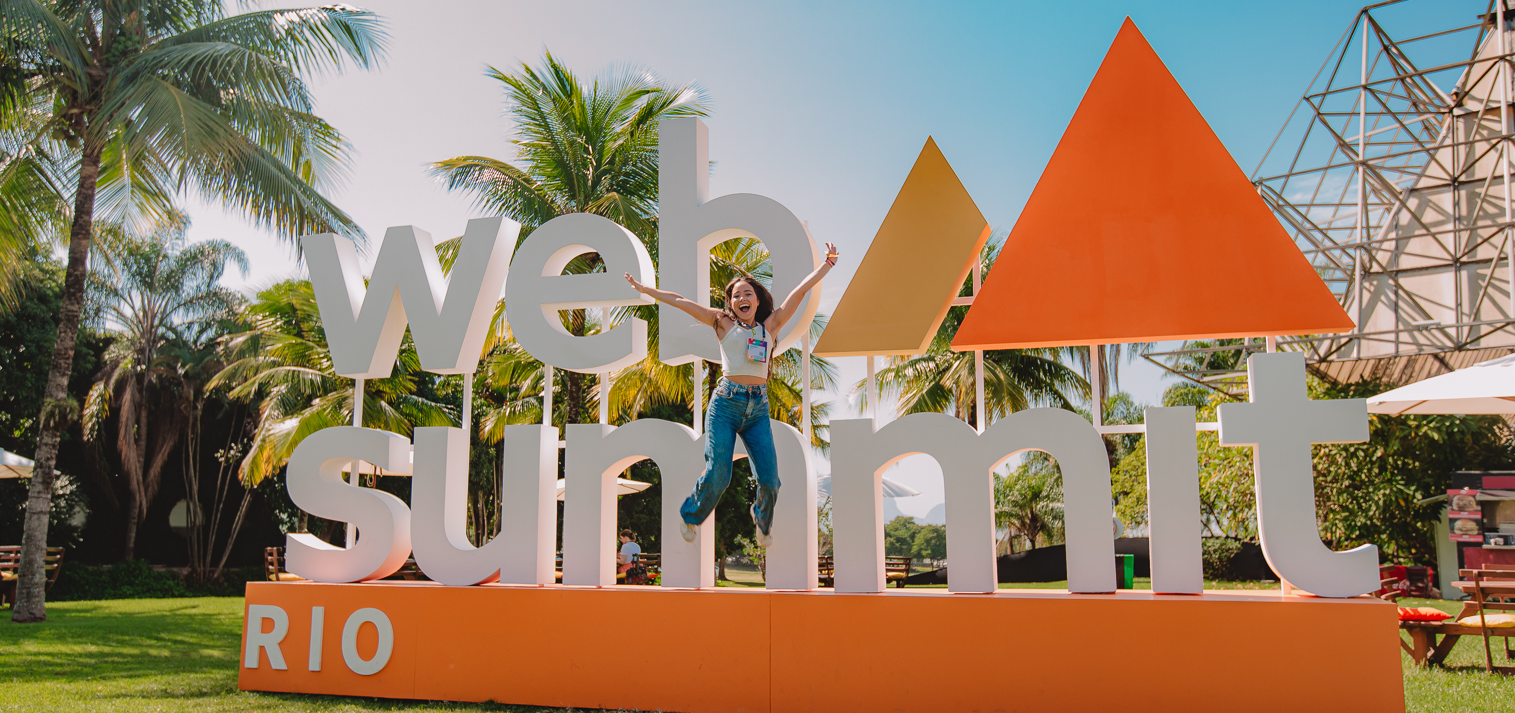 Web Summit Rio attendee leaps in front of a Web Summit Rio sign, on a bright day. There are palm trees visible in the background.