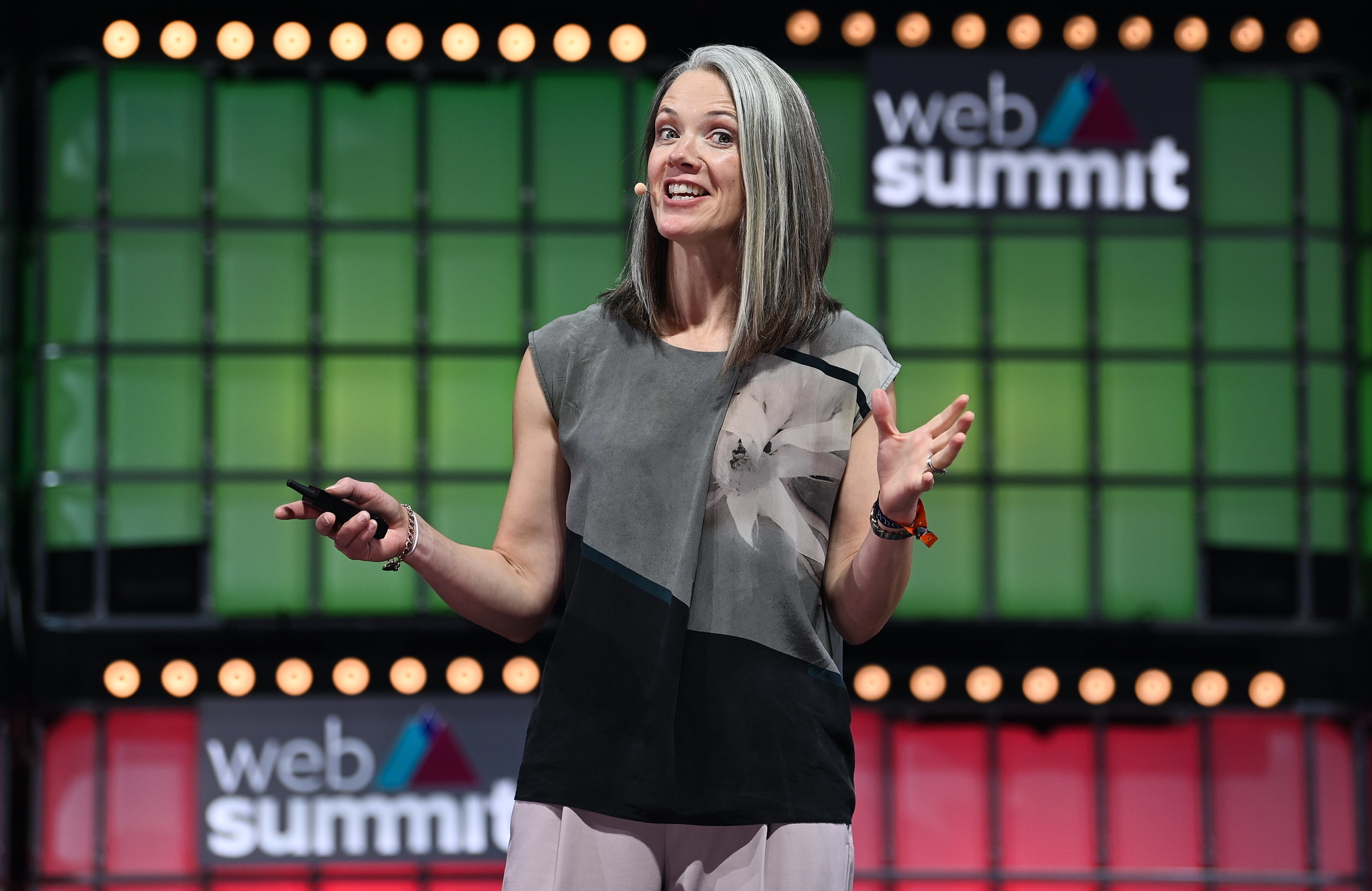 Photograph of Alison Darcy, founder and president of Woebot Health, speaking on stage at Web Summit. Alison is gesturing with hands and appears to be holding a remote. Alison is wearing an on-ear microphone and appears to be speaking. There are two Web Summit logos visible in the background.