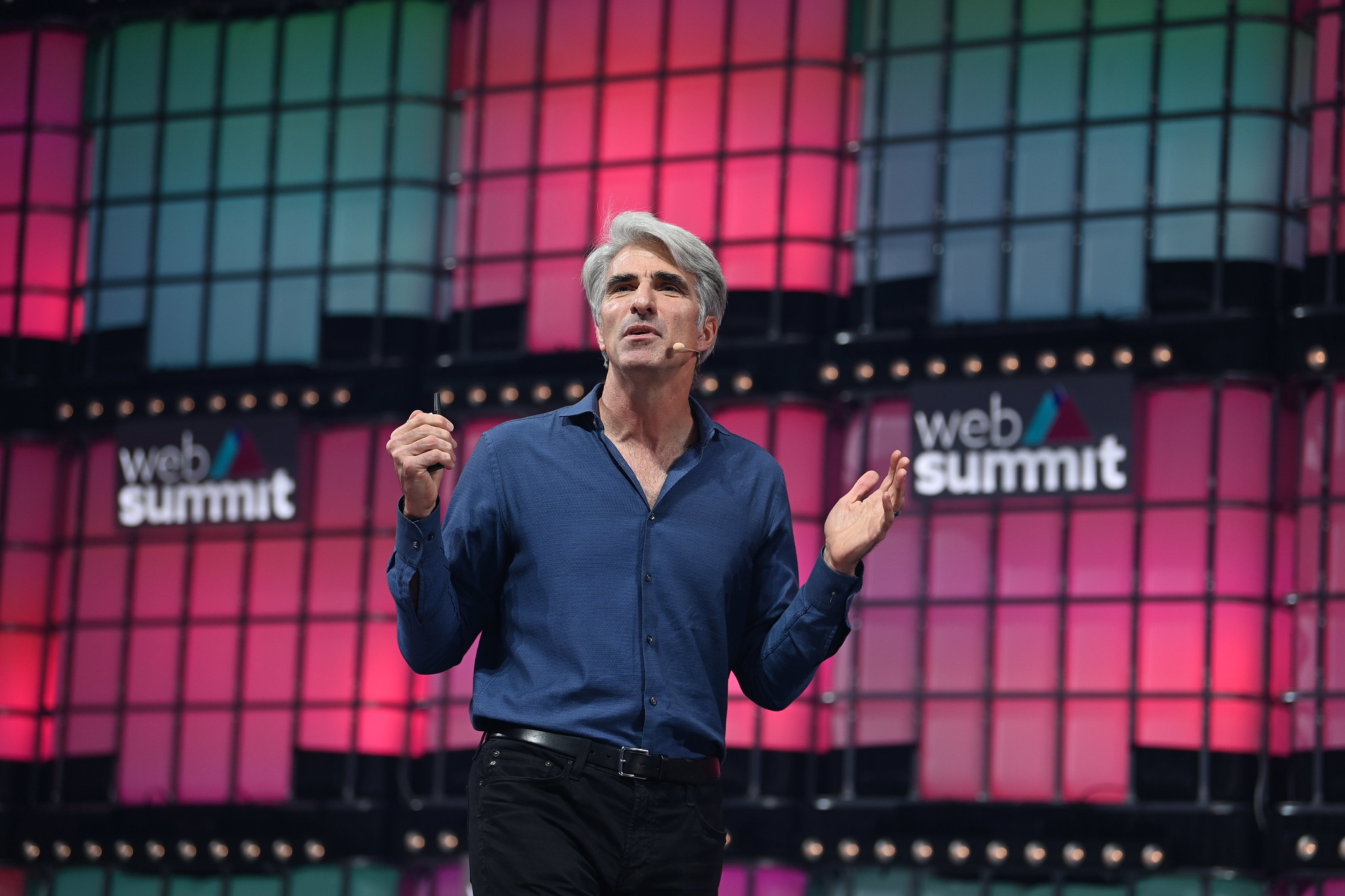 Photograph of a person (Craig Federighi, senior VP of software engineering at Apple) speaking on stage at Web Summit. They are gesturing with their hands and wearing an on-ear microphone.