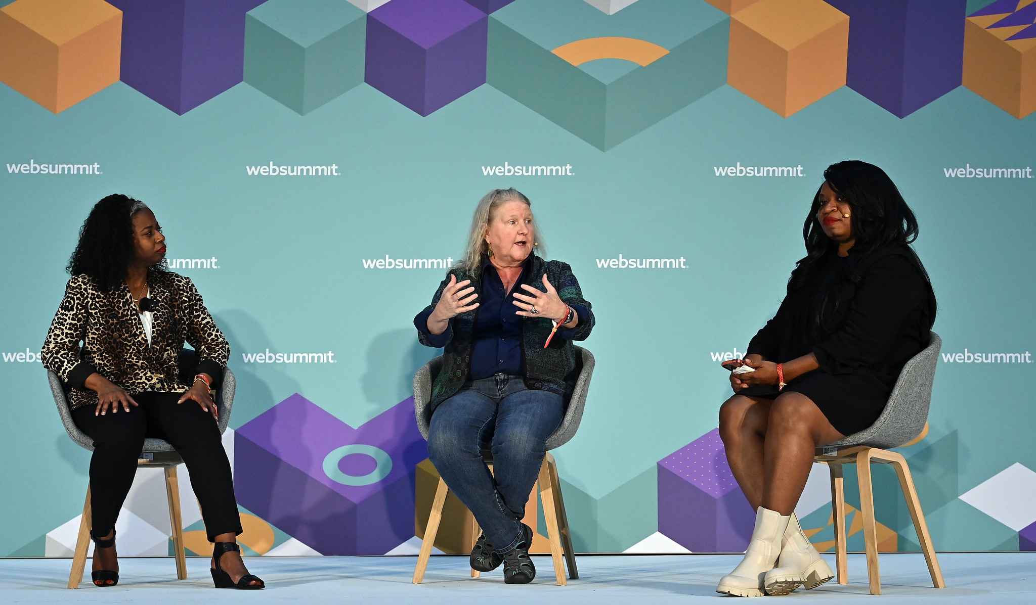 A photograph of three people speaking on stage at Web Summit. They are seated and appear to be having a discussion.