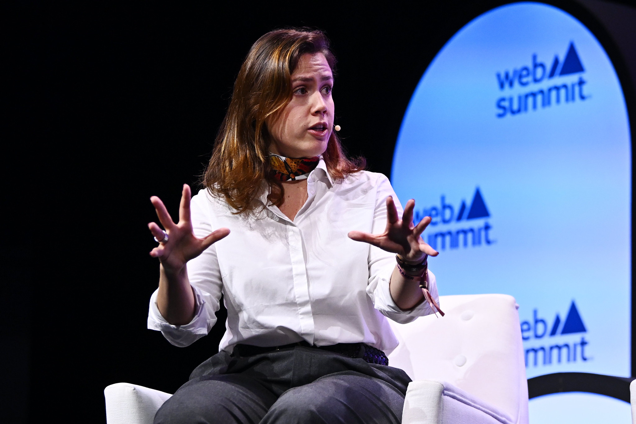 Photograph of Lidia Pereira, member of the European parliament, speaking on stage at Web Summit. Lidia is sitting on a chair and using hands to gesture emphatically while speaking. Lidia is wearing an on-ear microphone and appears to be speaking to another person.