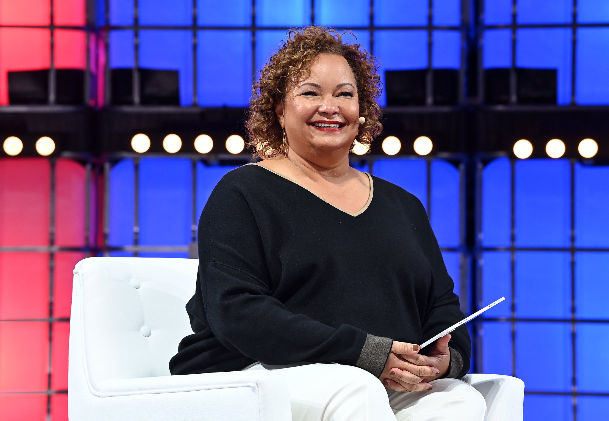 A photograph of Lisa Jackson, VP of environment, policy and social initiatives at Apple, speaking on stage at Web Summit. Lisa is sitting on a chair and appears to be holding a tablet or piece of paper. Lisa is smiling, and is wearing an on-ear microphone.
