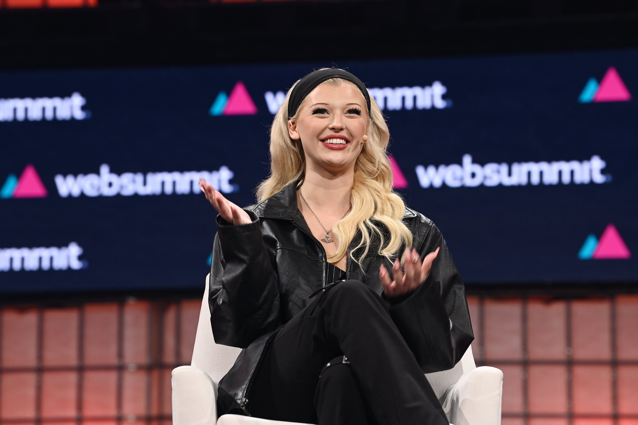 A photograph of Loren Gray, social media personality and songwriter, speaking on Centre Stage at Web Summit. Loren appears to be speaking to a crowd, and is sitting on a chair. Loren is gesturing with hands. The Web Summit logo is visible in the background.