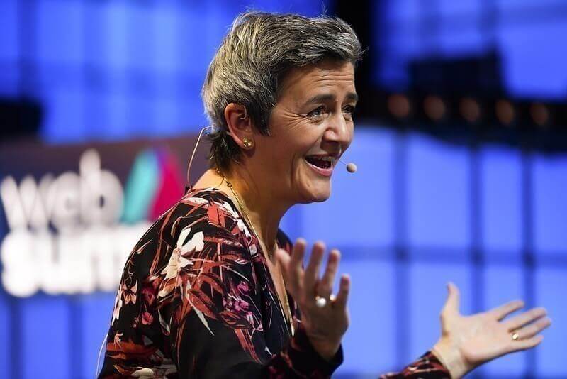 A photograph of Margrethe Vestager, the European Commissioner for Competition, speaking on stage at Web Summit. Margrethe is facing away from the camera and appears to be wearing an on-ear microphone. Margrethe appears to be speaking.