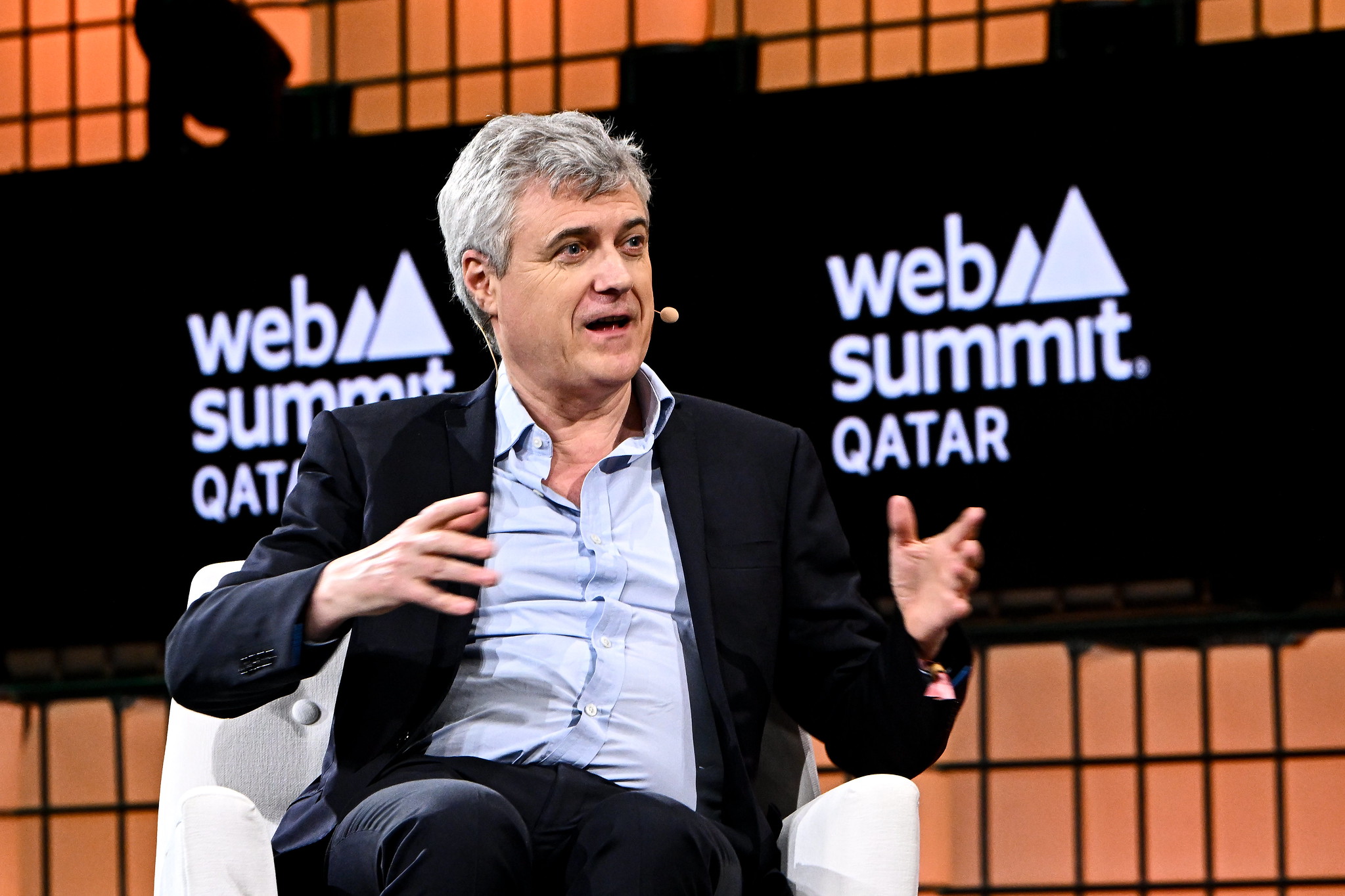 A photograph of Mark Read, CEO of WPP, speaking on stage at Web Summit Qatar. Mark is gesturing with hands and wearing an on-ear microphone. Mark is sitting on a chair and appears to be speaking. The Web Summit Qatar logo is visible in the background.