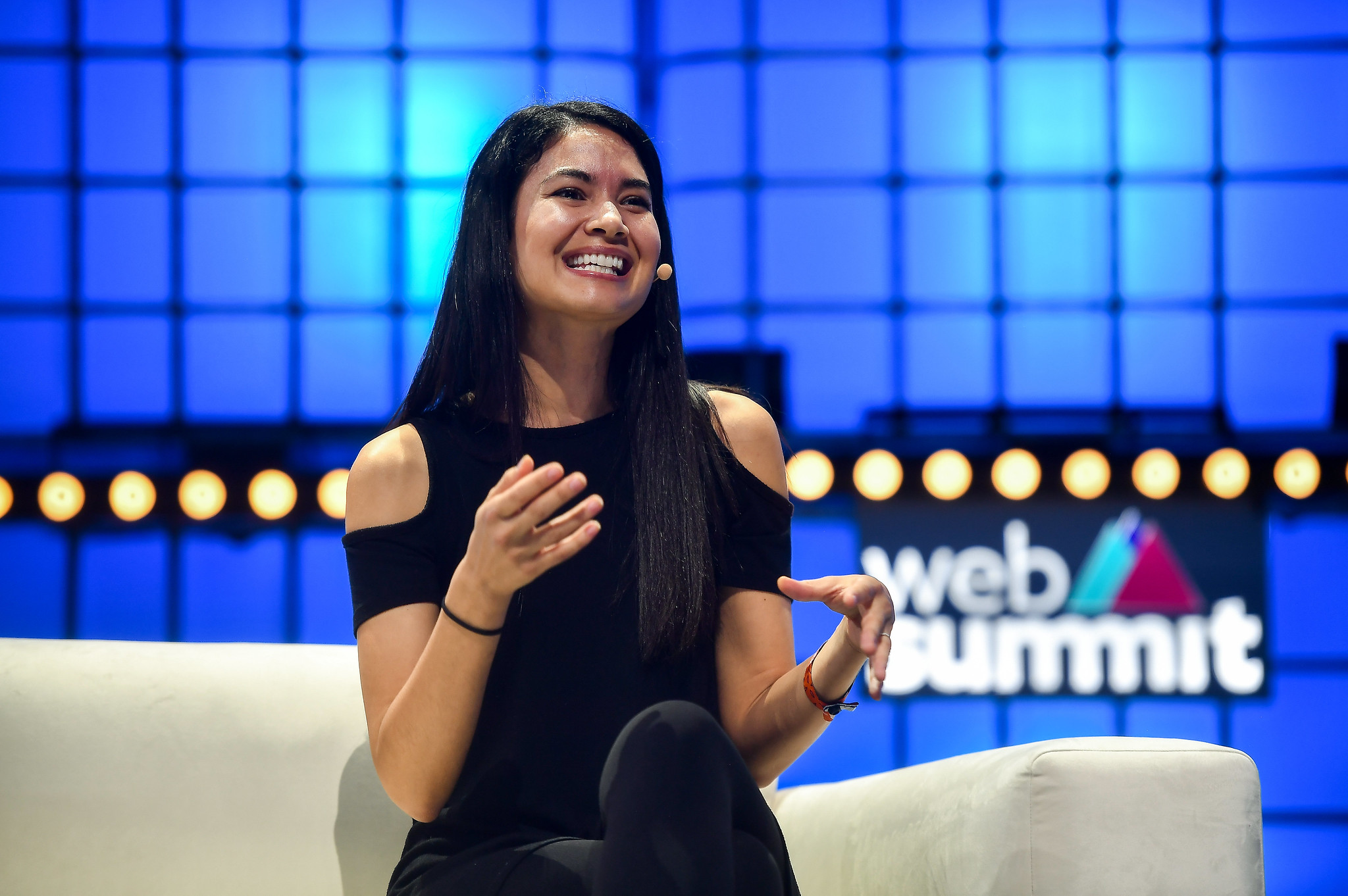 A photograph of Melanie Perkins, co-founder and CEO of Canva, speaking on stage at Web Summit. Melanie is sitting on a chair and appears to be speaking. Melanie is smiling, and wearing an on-ear microphone. The Web Summit logo is visible in the background.