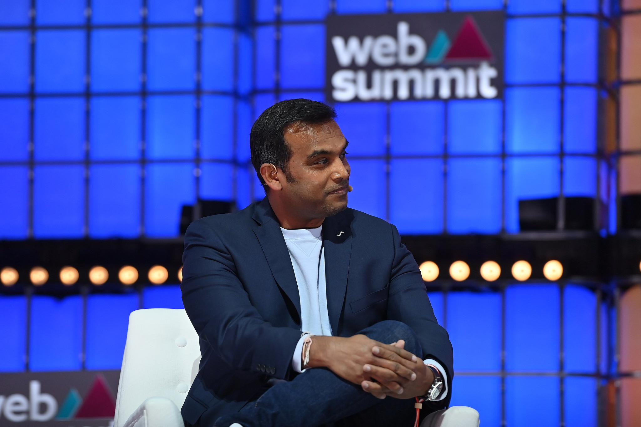 A photograph of a person (Parag Parekh, Chief Digital Officer of IKEA) sitting on a chair on stage at Web Summit. They have their hands crossed and placed on their knee. They appear to be engaged in conversation. The Web Summit logo is visible in the background.