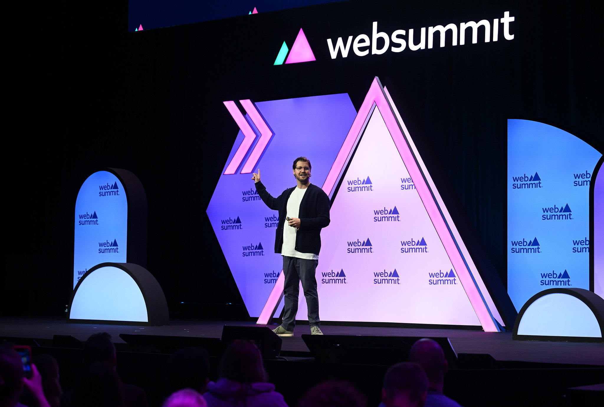 A photograph of Stevie Johnson, managing director of Disrupt, speaking on stage at Web Summit 2023. The photograph is zoomed-out, and there appears to be a crowd of people sitting at the stage. Stevie is standing on stage and appears to be speaking to the audience. The Web Summit logo appears above the stage.
