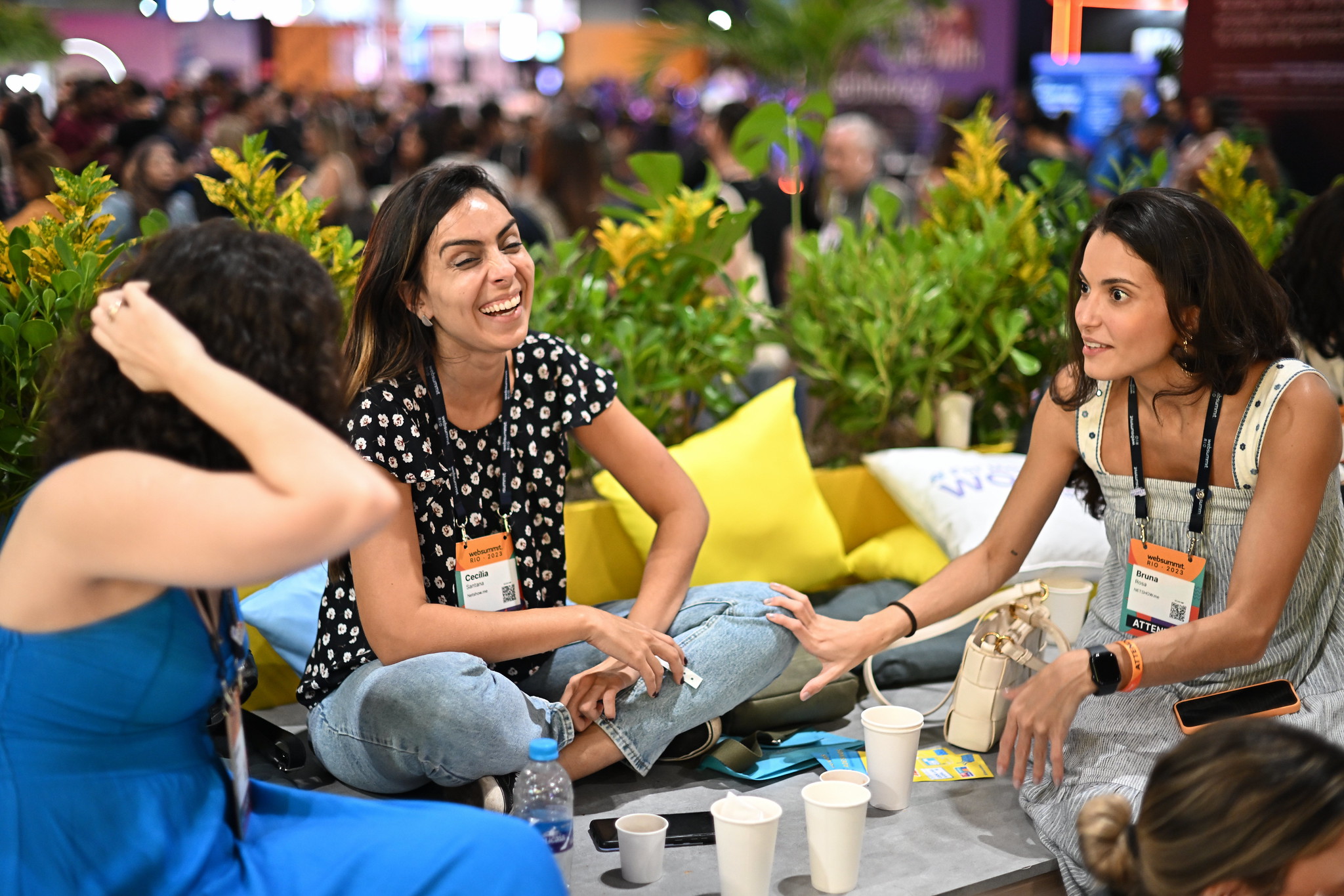 Three people sit on a low table. They appear to be speaking. One person has their back to the camera. They're scratching their head. Another person is sitting among cushions with their legs crossed and their forearms resting on their legs. They are smiling. The third person is resting their hand on the second person's knee. They appear to be engrossed in conversation.