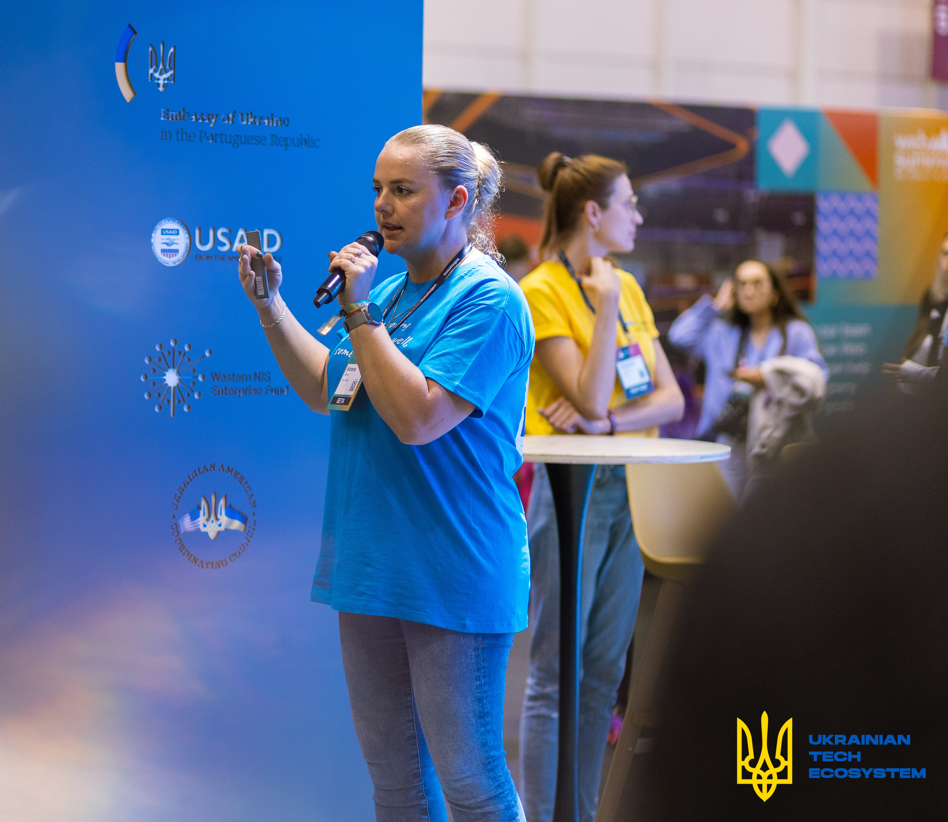 A photograph of a person speaking into a microphone at Web Summit. The person is holding a microphone in their left hand and appears to be gesturing with their right hand.