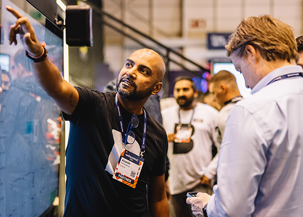 A photograph of a Web Summit attendee pointing in the air. Another attendee stands in front. The person pointing is wearing a lanyard.