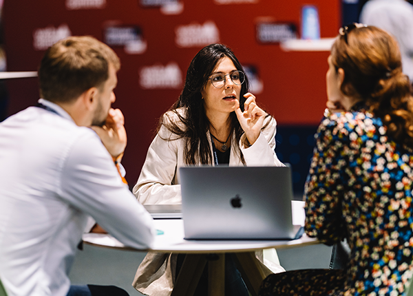 A photograph of three people sitting at a roundtable. One person appears to be talking and the other two are listening, with their backs faced to the camera. There is an Apple MacBook sitting on the table between the three people.