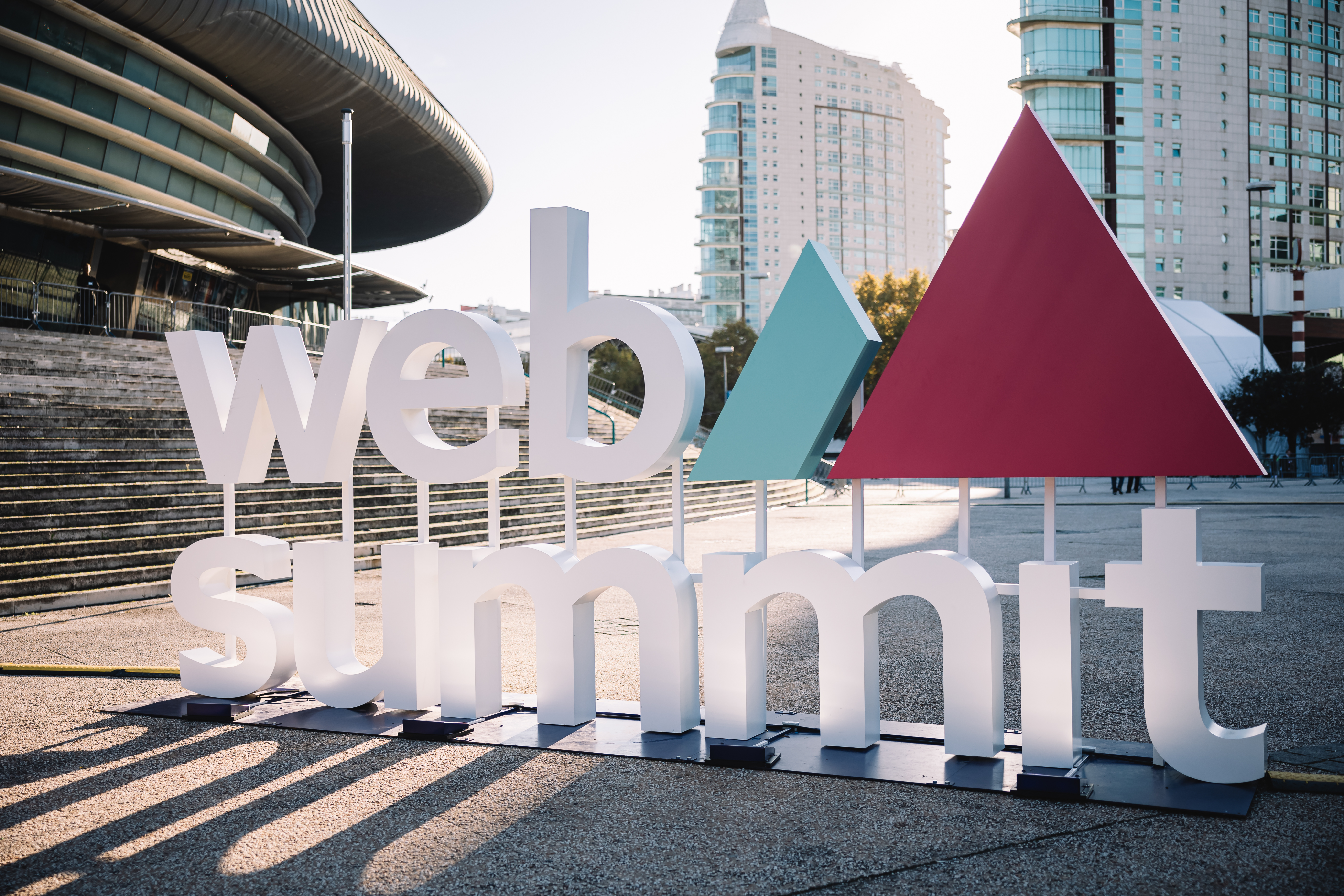 A photograph of a freestanding Web Summit sign and logo outside of the event. There are high-rise buildings visible in the background.