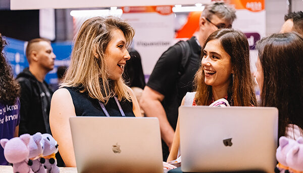 A photograph of two attendees facing each other. They each have a laptop in front of them. They are smiling at eachother. In the background, there are other attendees walking past.